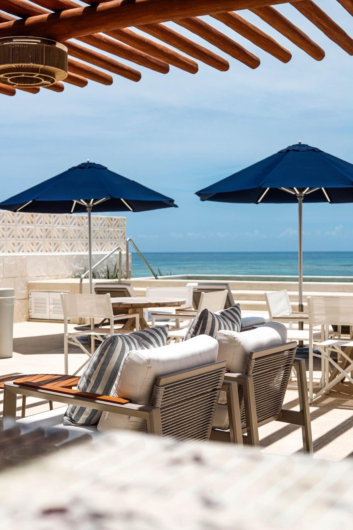 Un salon de plage avec des sièges en plein air, deux parasols et une vue sur l’océan.