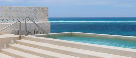 A pool with steps leading to the water, a clear view of the ocean, and a patterned fence in the background.