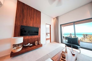A modern living room with a flat-screen TV, wooden paneling, a large window with a view of the ocean, and a seating area with a table. - Apt Jaguar: Balcony with Sea View & Rooftop Pool (Puerto Morelos)