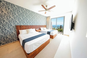 A hotel room with two beds, a ceiling fan, a large window with a view, and a TV mounted on the wall. - Apt Jaguar: Balcony with Sea View & Rooftop Pool (Puerto Morelos)