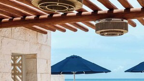 Un área de descanso al aire libre con vista al mar, que incluye sillones, sombrillas y una pérgola con luces colgantes.