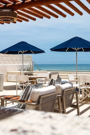 A beachside lounge with outdoor seating, two umbrellas, and a view of the ocean. - Apt Flamingo: Relaxing Getaway with Shared Pool (Puerto Morelos)