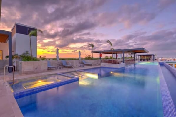 A swimming pool with lounge chairs and a covered seating area, surrounded by a modern building.