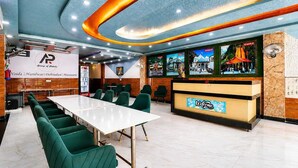Uma sala de conferências com uma mesa comprida e cadeiras verdes, uma recepcionista com uma placa de 'Welcome' e uma parede com quadros emoldurados.