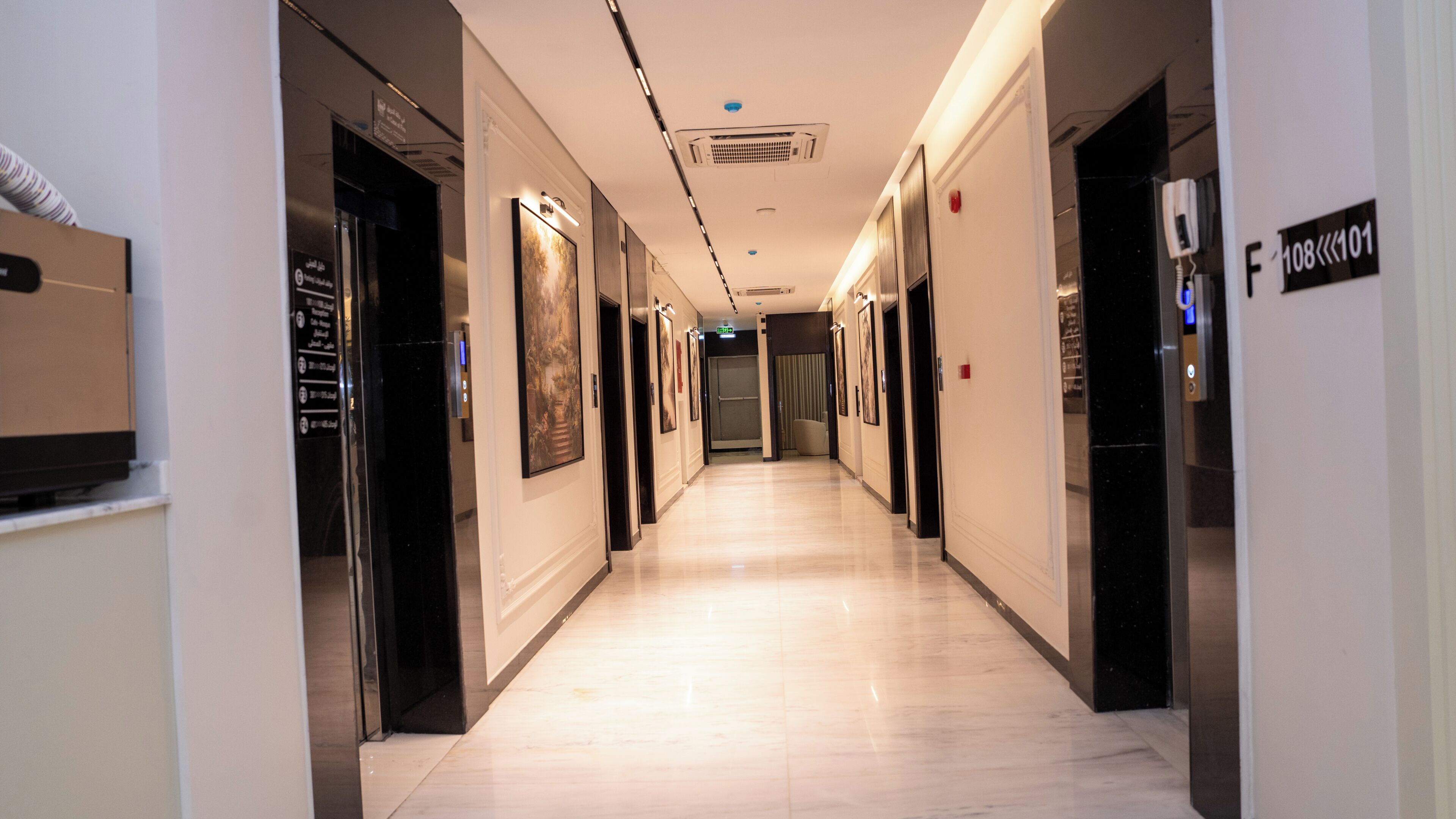 A hotel corridor with a marble floor, framed artwork on the walls, and an elevator on the right.