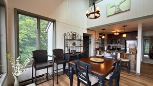Comedor con mesa y sillas de madera, un candelabro y vistas al exterior a través de amplias ventanas.
