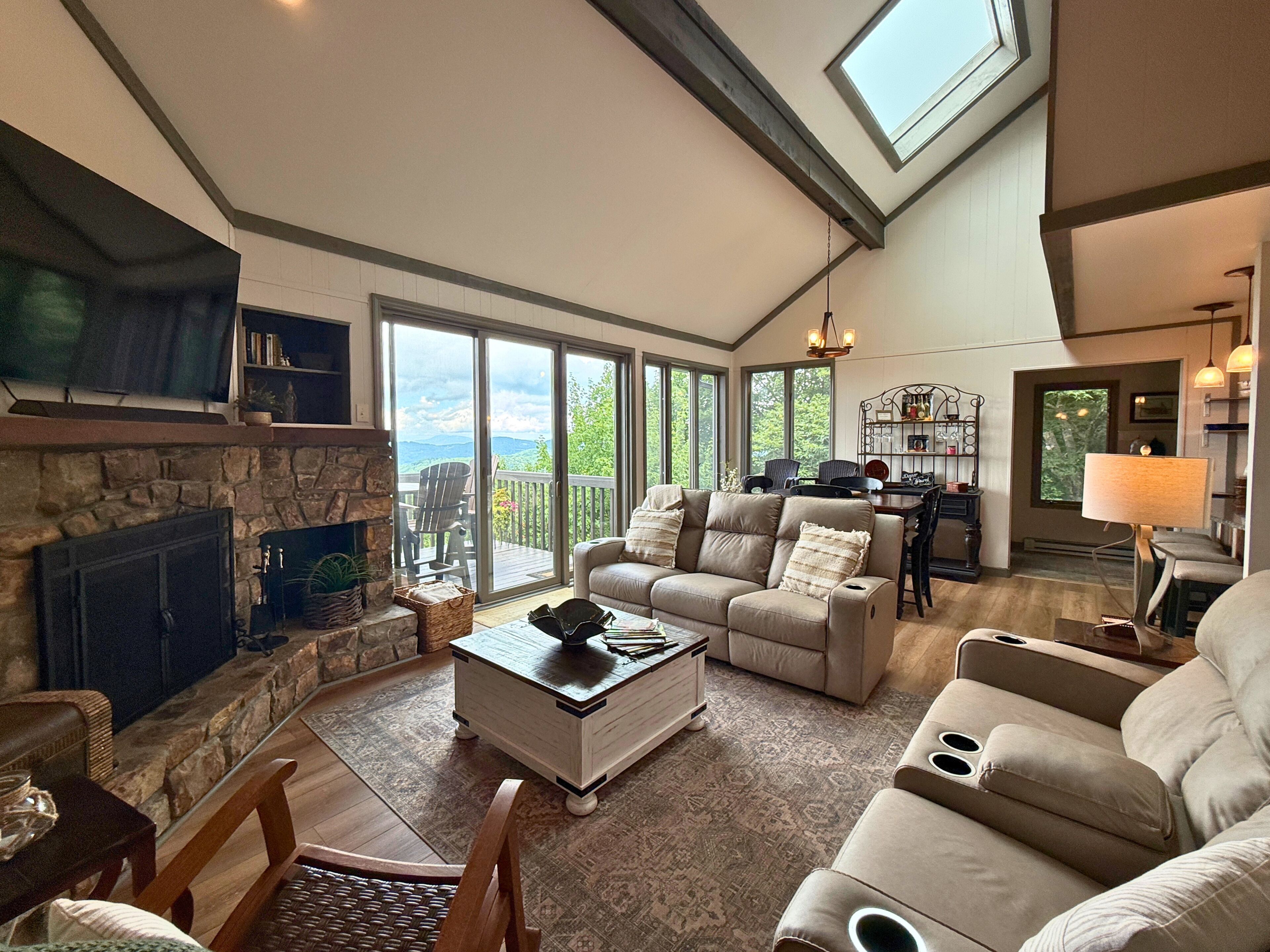 A spacious living room with a stone fireplace, comfortable seating, a dining area, and a large sliding glass door leading to a deck with a view.