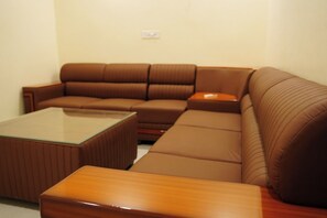 A brown leather sofa with a glass coffee table and a wooden side table in a room with plain walls. - The Gautam Residency (Jaunpur)