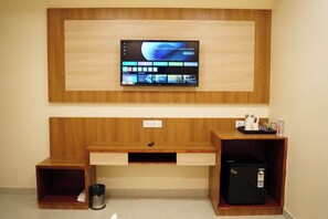 A hotel room with a TV mounted on a wooden wall, a wooden desk with a trash bin, and a small black refrigerator.