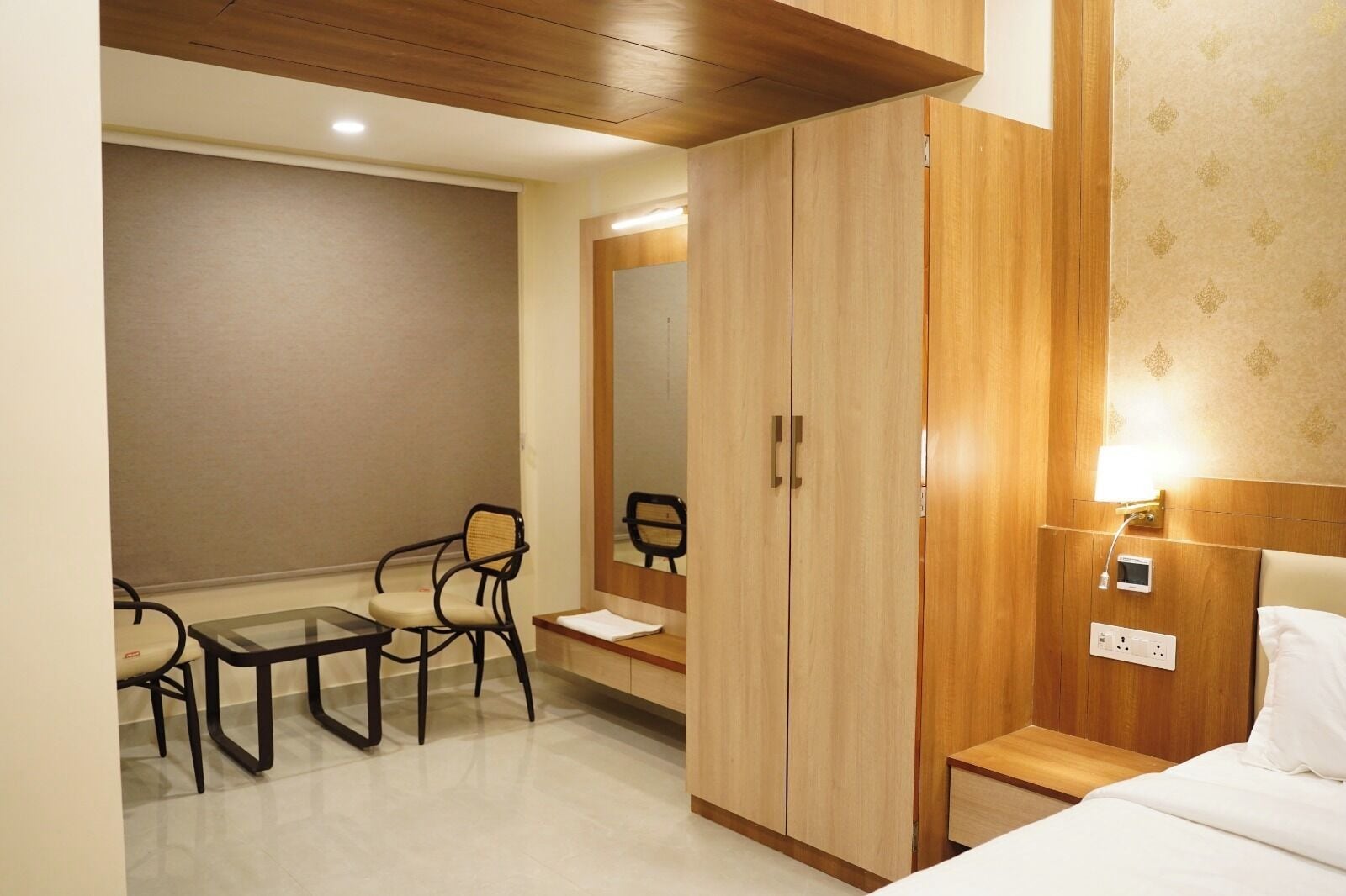 A hotel room with a bed, a desk with a chair, a wardrobe, and a bedside table with a lamp.