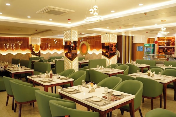 A restaurant with green upholstered booths and tables, modern lighting fixtures, and a bar area in the background.