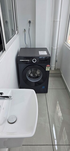 A compact laundry area with a Bosch washing machine, a white sink, and tiled flooring. - Lovely 2-bedroom apartment with AC for a relaxing stay (Ernakulam)