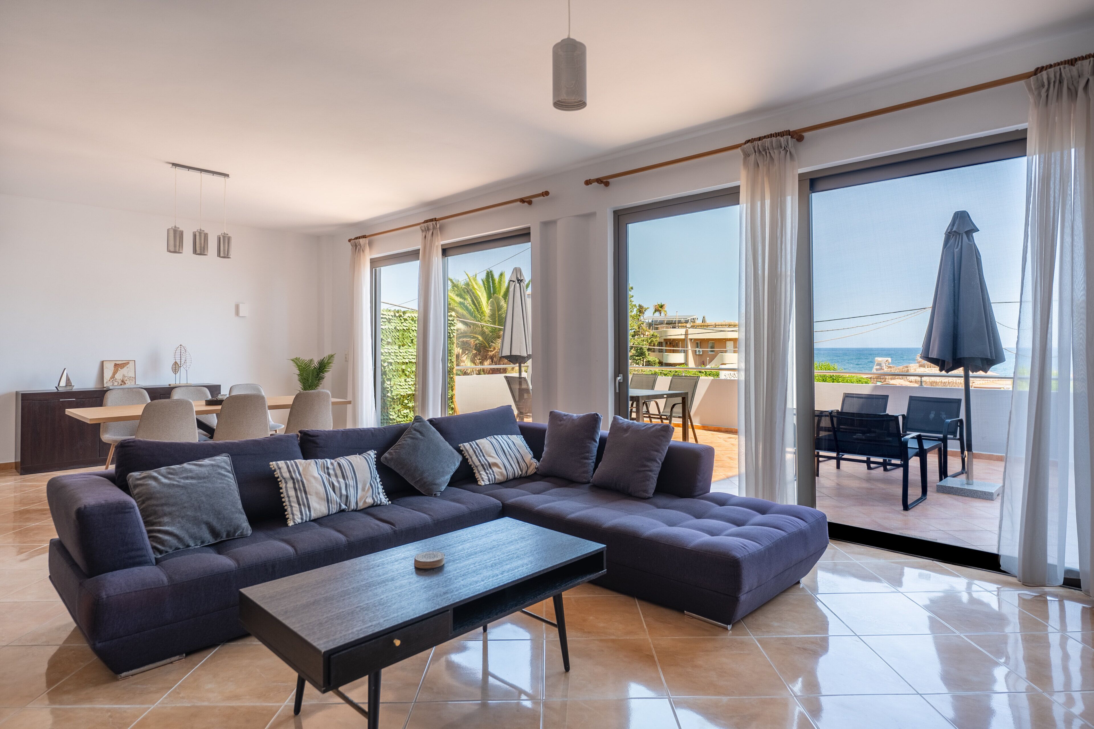 A modern living room with a sectional sofa, a coffee table, and a dining area with chairs and a table. Large windows offer a view of the outdoors.