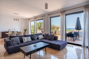 A modern living room with a sectional sofa, a coffee table, and a dining area with chairs and a table. Large windows offer a view of the outdoors. - Seaside House (Galatas)