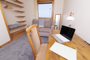 Een kamer met een houten bureau, een laptop, een notitieboek, een stoel, een bank, een raam en een plank met manden.
