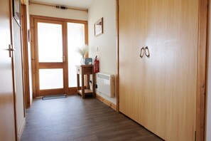 Een gang met een houten deur, een radiator, een klein tafeltje met een vaas en een brandblusser.