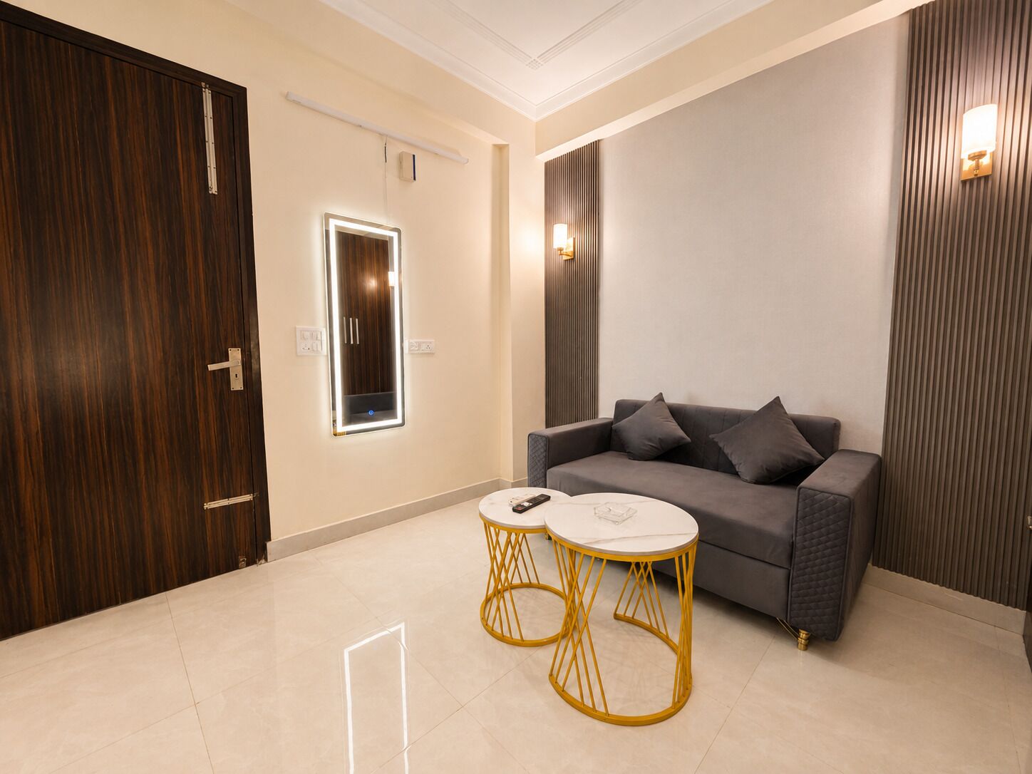 Uma sala de estar moderna com um sofá cinzento, duas mesas redondas pequenas com pés amarelos e um espelho grande com iluminação embutida.