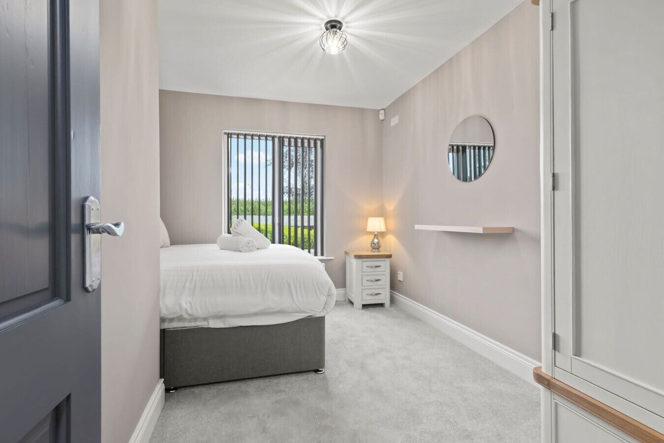 Een slaapkamer met een bed, nachtkastje, lamp en uitzicht door het raam.
