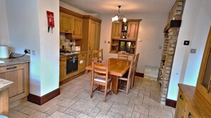 A kitchen with wooden cabinets, a dining table, and chairs. There is a stone fireplace and a hanging light fixture. - Thorn Cottage by Escape Ordinary (Ballycastle)