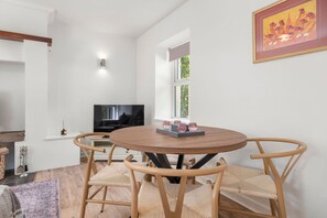 Un coin repas avec une table ronde en bois, quatre chaises en osier, un téléviseur posé sur un meuble et une fenêtre donnant sur un paysage verdoyant.