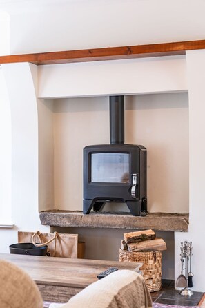 A wood-burning stove in a room with a stone hearth, a woven basket, and a remote control on a wooden table. - Horseshoe Cottage by Escape Ordinary (Derrygonnelly)