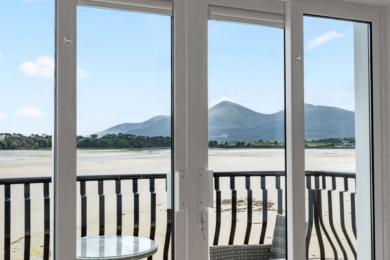 A view from a room with a glass door leading to a balcony overlooking a beach and mountains.