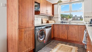 A modern kitchen with wooden cabinets, a built-in washing machine, and a window with a view of the outside. - The Lookout · Dundrum Bay · Balcony with Bay Views (Dundrum)