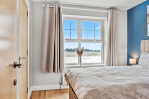 A bedroom with a large window, a bed with a white bedspread, and a vase with dried flowers on the windowsill. - The Lookout · Dundrum Bay · Balcony with Bay Views (Dundrum)