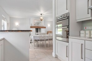 A modern kitchen with white cabinetry, a built-in oven, and a dining area with a table and chairs. - Orchard View Cottage | Escape Ordinary (Derrygonnelly)