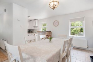 A modern kitchen with white cabinets, a dining table with chairs, a clock, and a window with a view of greenery. - Orchard View Cottage | Escape Ordinary (Derrygonnelly)