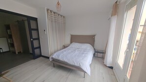 Ein Schlafzimmer mit einem Bett, einer Deckenleuchte, einer Tür und Blick durch ein Fenster.