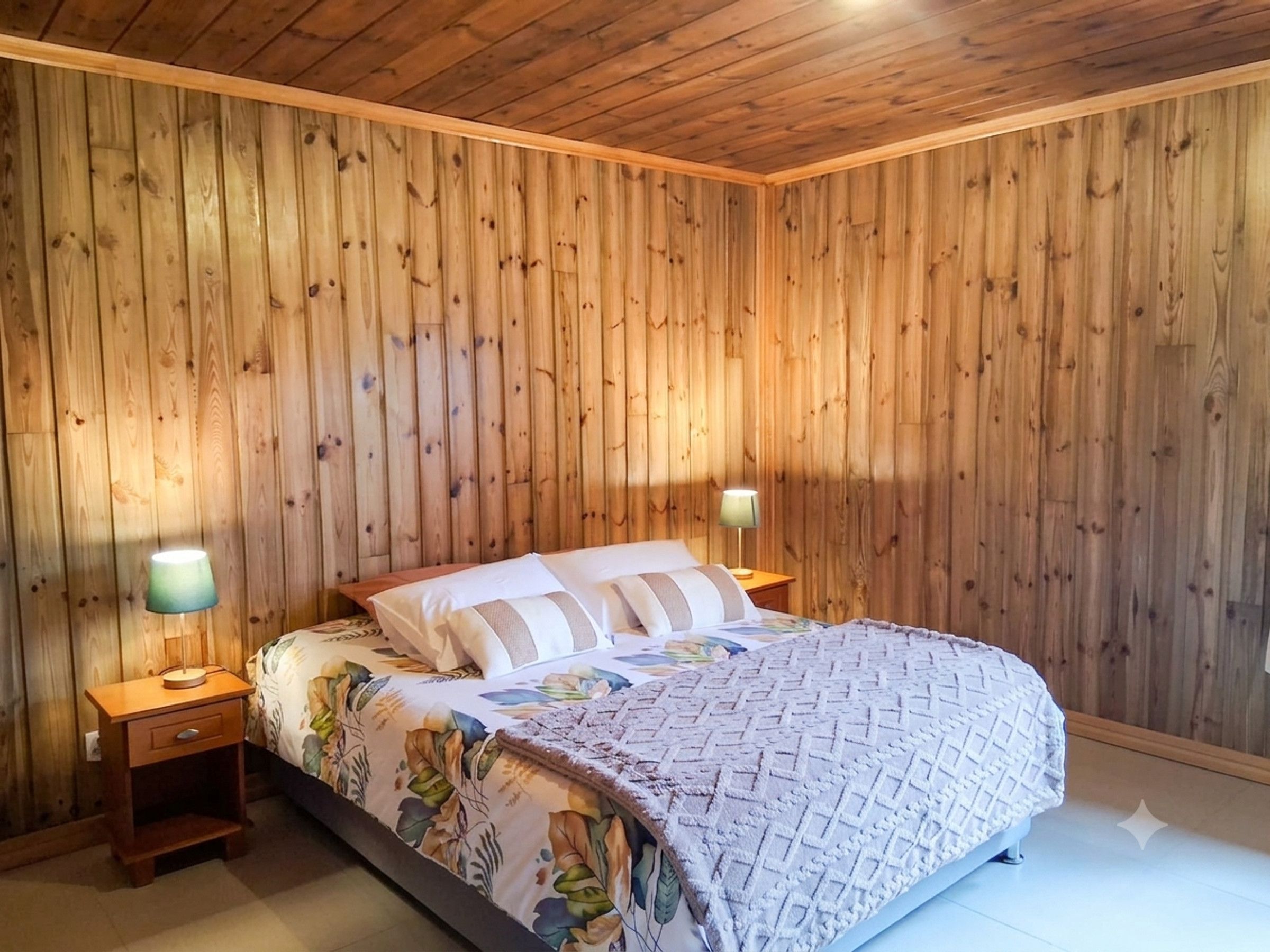 Ein Schlafzimmer mit einer holzverkleideten Wand, einem Bett mit floralem Überwurf, zwei Nachttischen mit Lampen und einem kleinen Holz-Nachttisch.