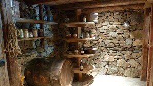 Una cocina rústica con paredes de piedra, estantes de madera, un barril grande y diversos utensilios de cocina.