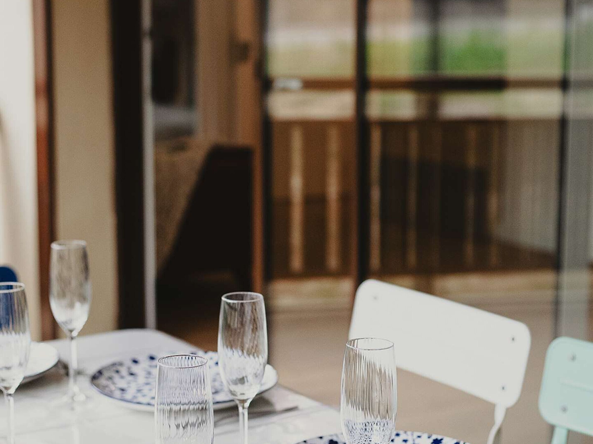 Una mesa dispuesta con cristalería transparente y platos, una silla blanca y vista a un balcón con baranda de madera.