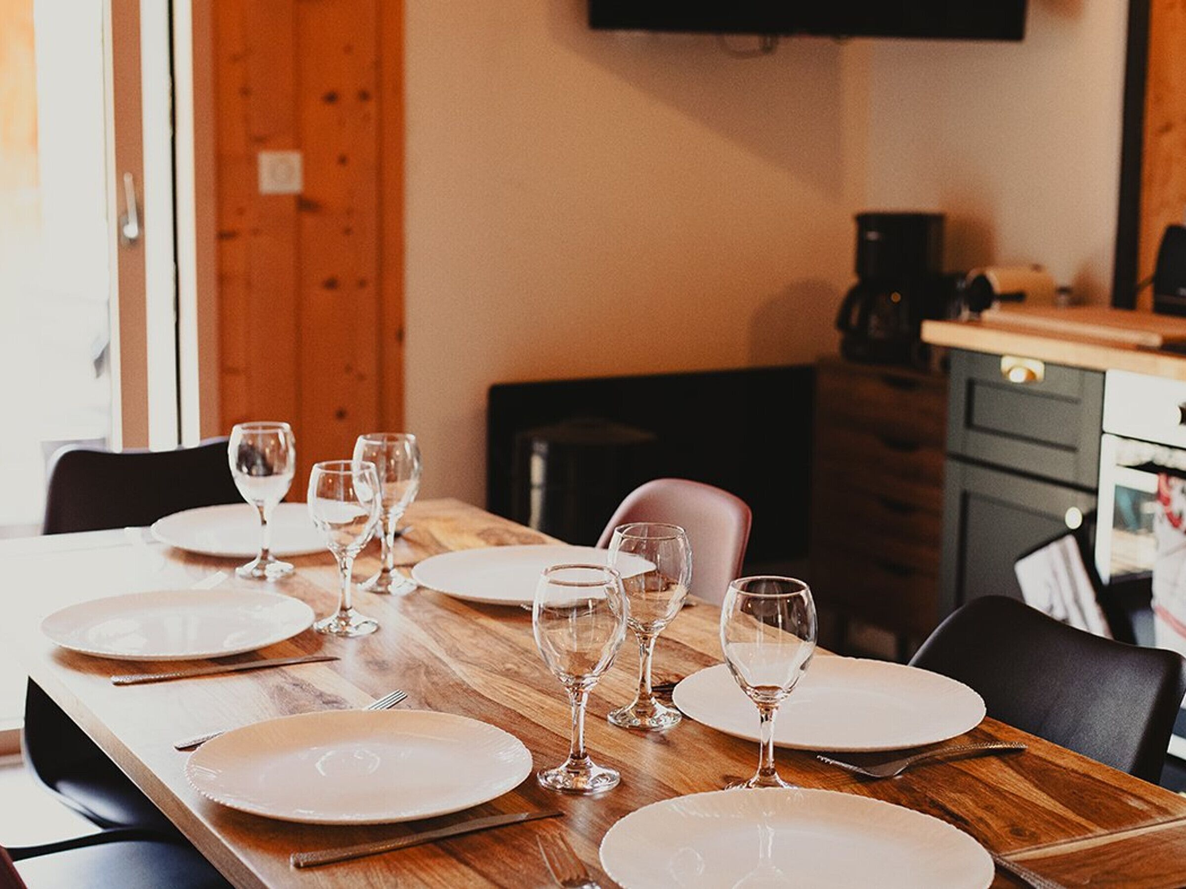 Una mesa de comedor dispuesta con platos, copas de vino, sillas de madera y un televisor montado en la pared.