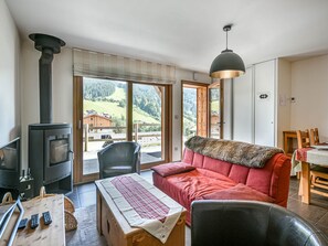 A cozy living room with a sofa, a table with remotes, a fireplace, and a view of the outdoors. - Charming T3 with parking - 5 guests (Les Gets)