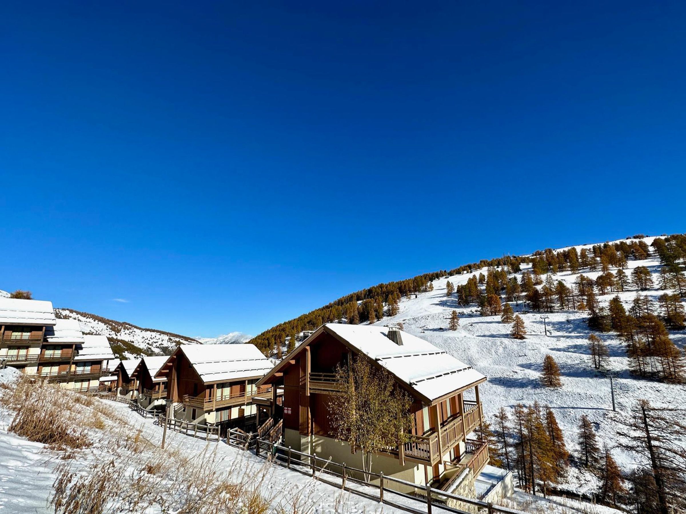 A snow-covered lodge with multiple wooden cabins, a ski lift, and a mountainous backdrop.