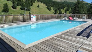 A rectangular swimming pool with a wooden deck and lounge chairs, surrounded by a wooden fence, set against a backdrop of green hills and a mountain. - Apartment pets, mountain view - 4 guests (Vars)