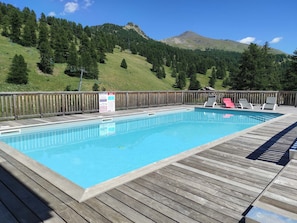 Ein rechteckiger Pool mit Holzterrasse und Liegestühlen, umgeben von einem Holzzaun, vor dem Hintergrund grüner Hügel und eines Berges.