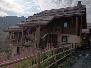 Eine Holzchalet mit Balkon und Treppe, umgeben von einem Holzzaun und blühenden Pflanzen.