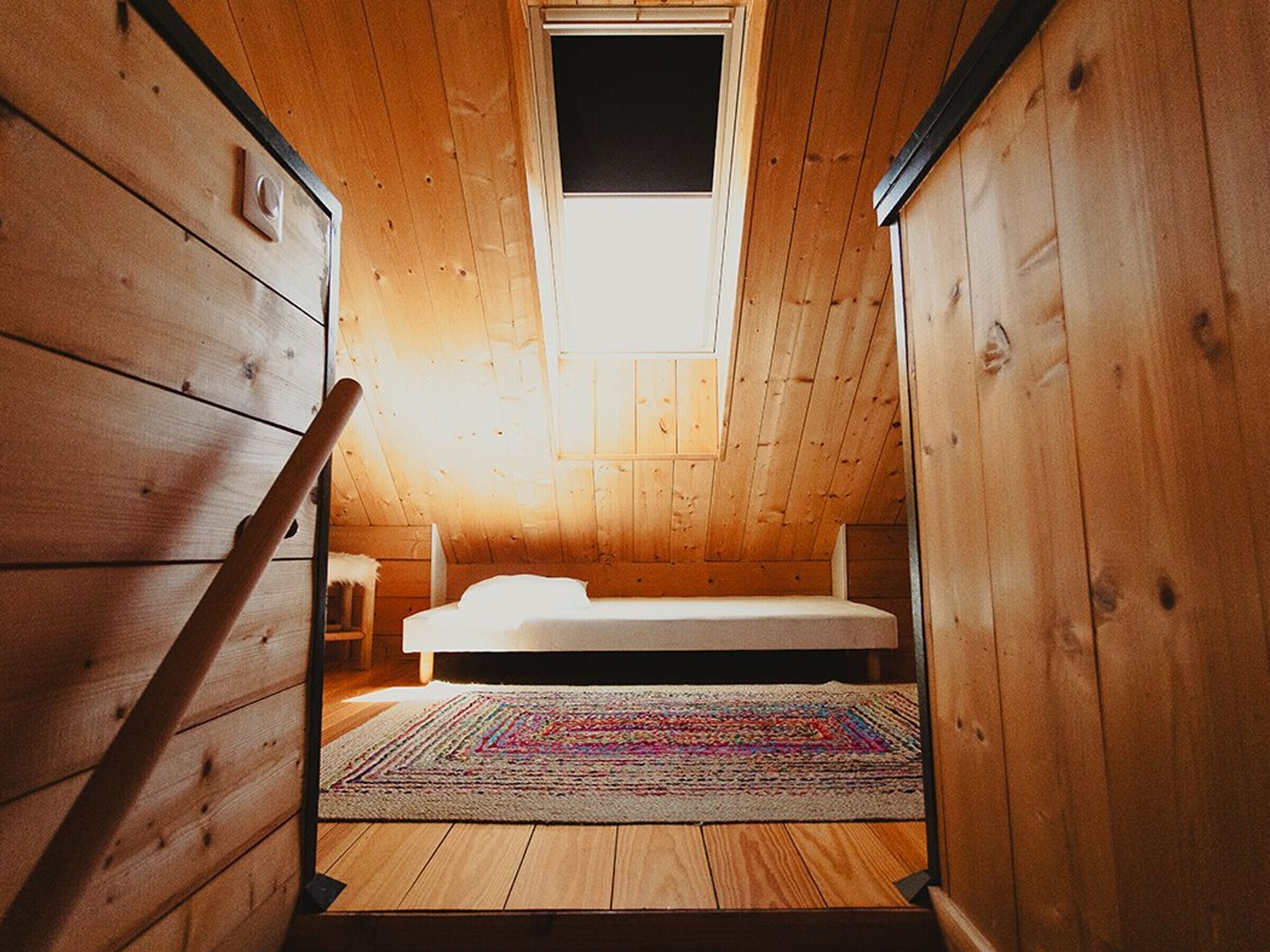 Een kleine kamer met houten wanden, een schuin dak, een enkel bed, een kleurrijk vloerkleed en een raam.