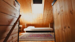 Ein kleiner, hölzerner Raum mit schrägem Dach, einem Einzelbett, einem bunten Teppich und einem Fenster.
