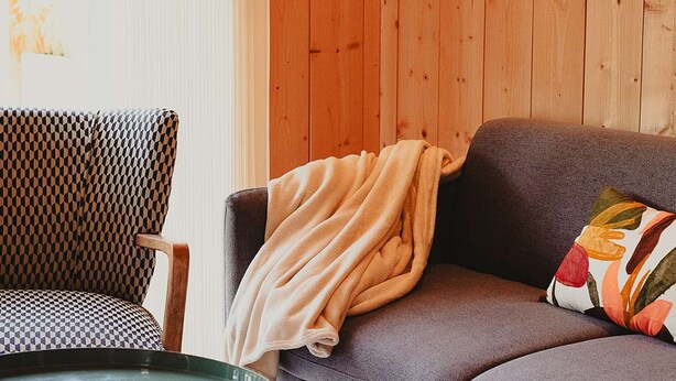 Ein Zimmer mit einer Holzwand, einem gemusterten Sessel, einem grauen Sofa mit Decke und einem Blumenkissen sowie einem Glastisch.