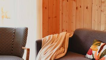 Een kamer met een houten wand, een gestreept fauteuil, een grijze bank met een deken en een bloemenkussen, en een glazen tafel.