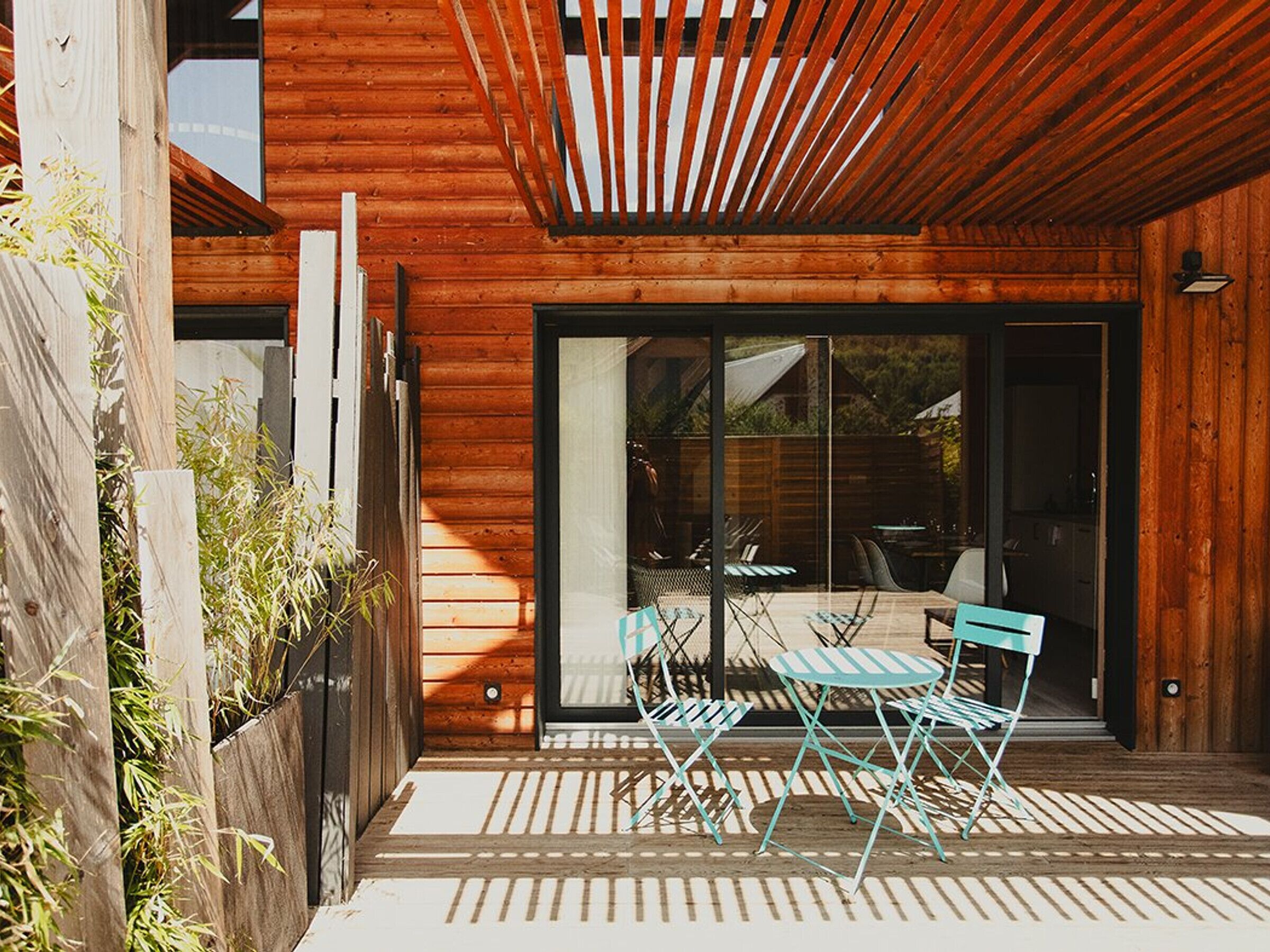 A modern outdoor patio with wooden flooring, a glass door leading to a dining area, and a metal railing.