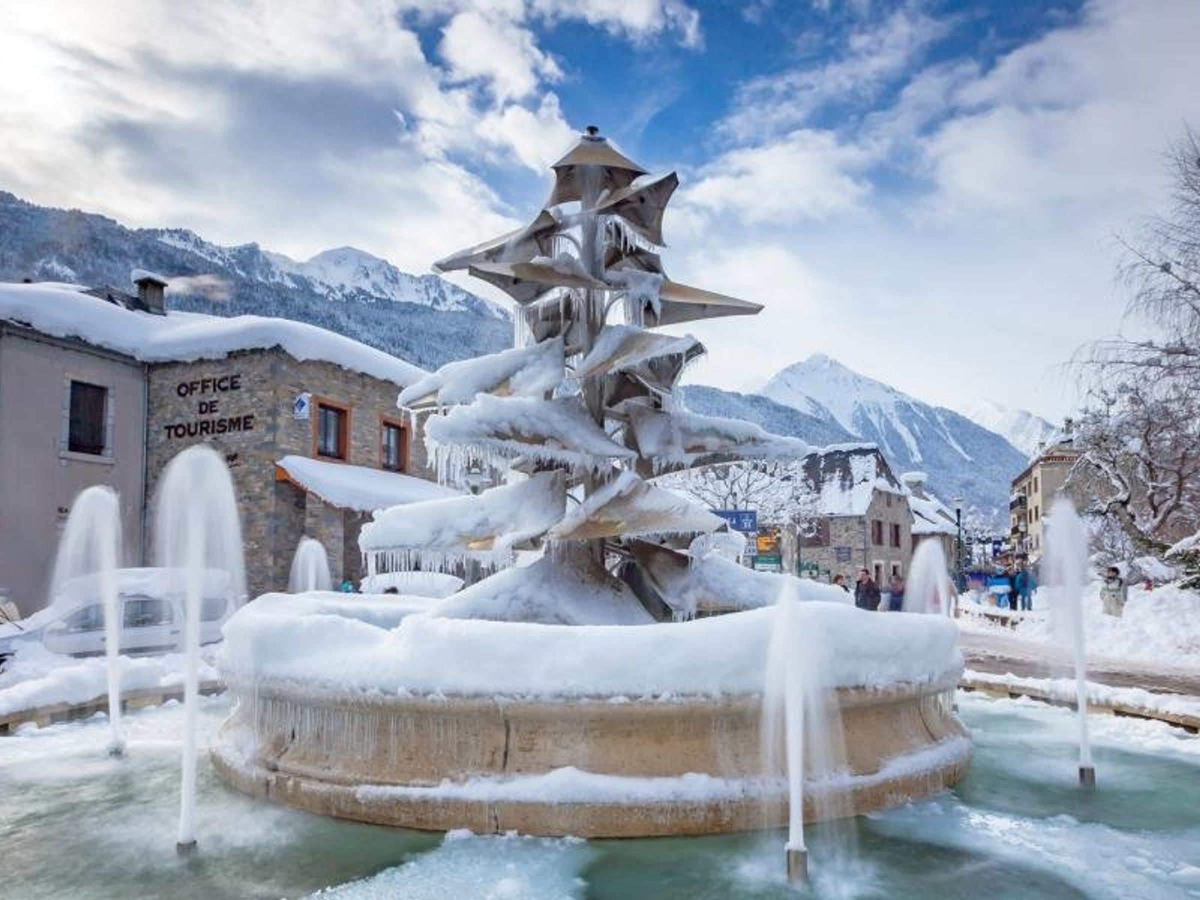 A snow-covered fountain with a mountainous backdrop, a stone building labeled 'Office de Tourisme,' and a clear sky.