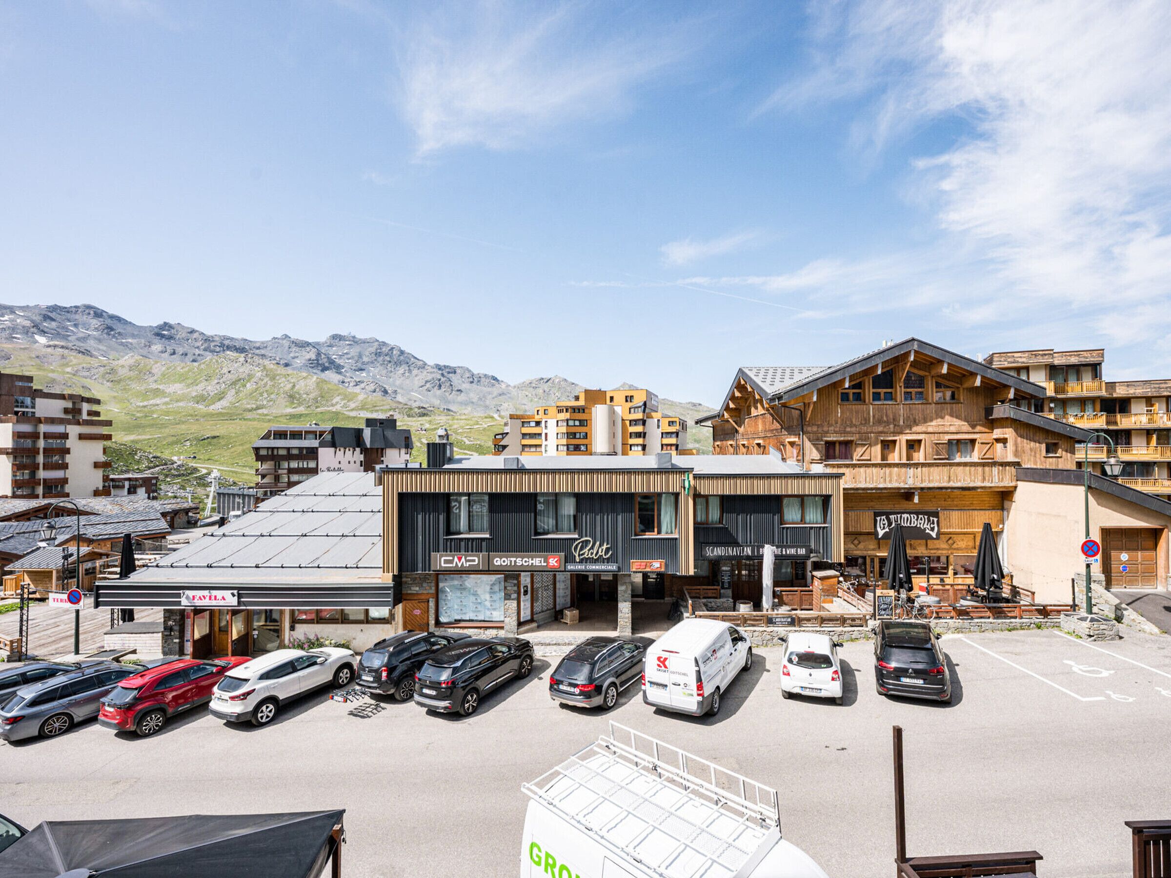 Un hotel con fachada moderna, un estacionamiento con varios coches y un paisaje montañoso de fondo.