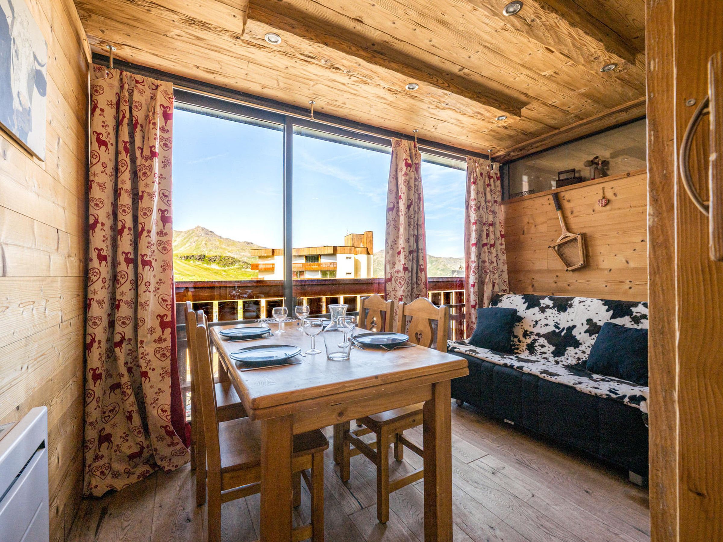 A wooden interior with a dining table set for four, a sofa, and a large window offering a view of a building and landscape.