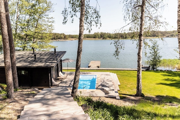 A wooden deck leading to a swimming pool with a view of a lake and a small building.