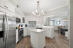 A modern kitchen with white cabinetry, stainless steel appliances, a central island, and a dining area with a table and chairs. - Windward Pointe 1204 by Liquid Life (Orange Beach)